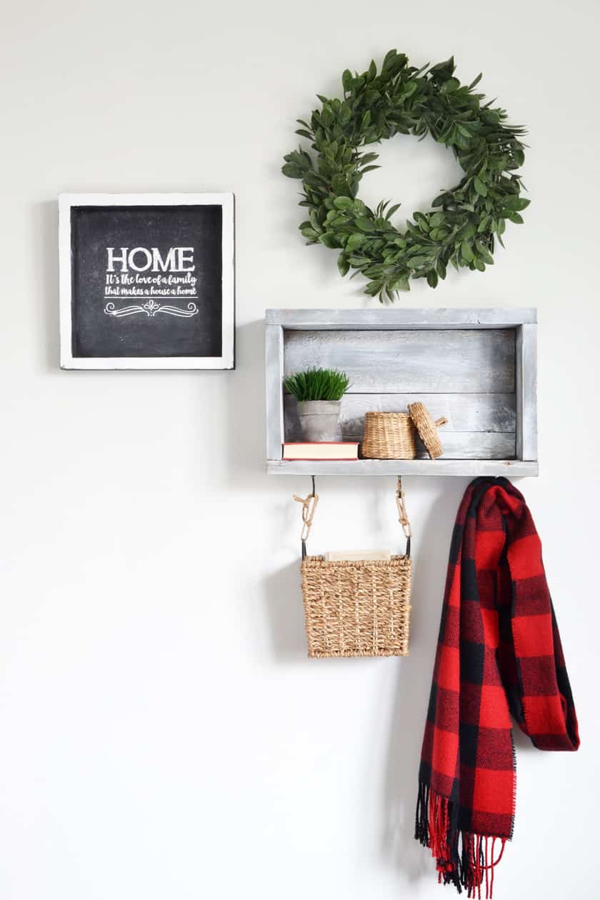 A rustic wall shelf with hooks holding a basket and winter scarf. A small basket is on the shelf with a plant beside it.