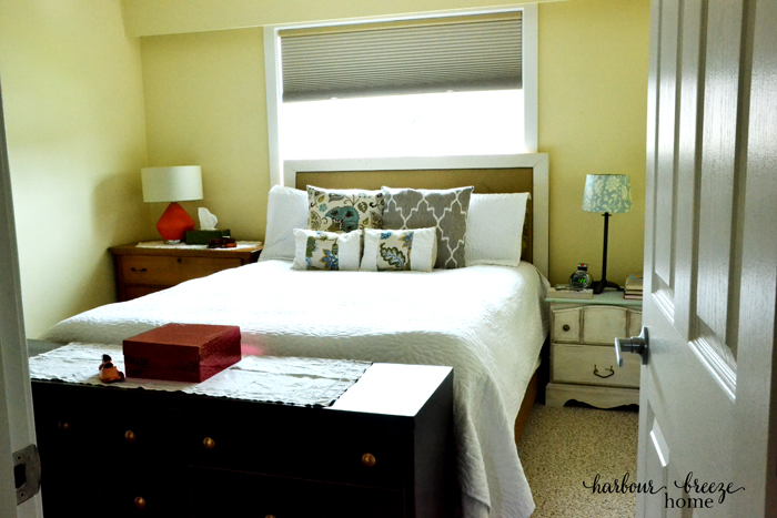 A queen bed under a window in the center of the main wall of a bedroom with a dresser on one side and a nightstand on the other. A long, low dresser sits at the foot of the bed.