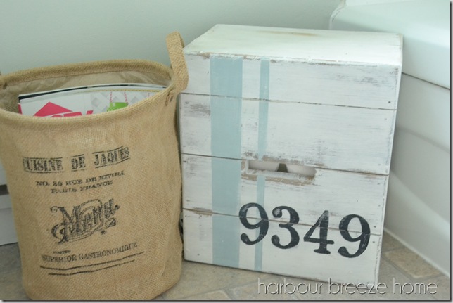 A large basket with magazines in it and a crate filled with toilet paper sitting on the floor in a bathroom.