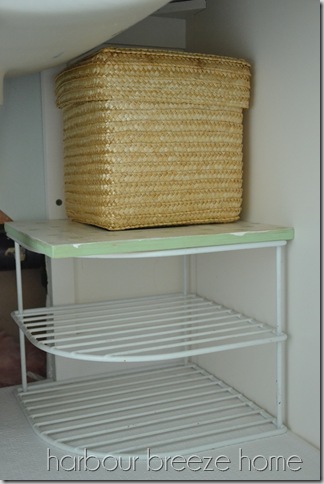 A wire corner kitchen shelf organizer in a bathroom vanity with a lidded basket on the top.