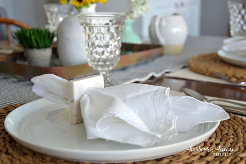 a white cloth napkin in a square napkin holder sitting on top of a white plate with a wicker charger underneath