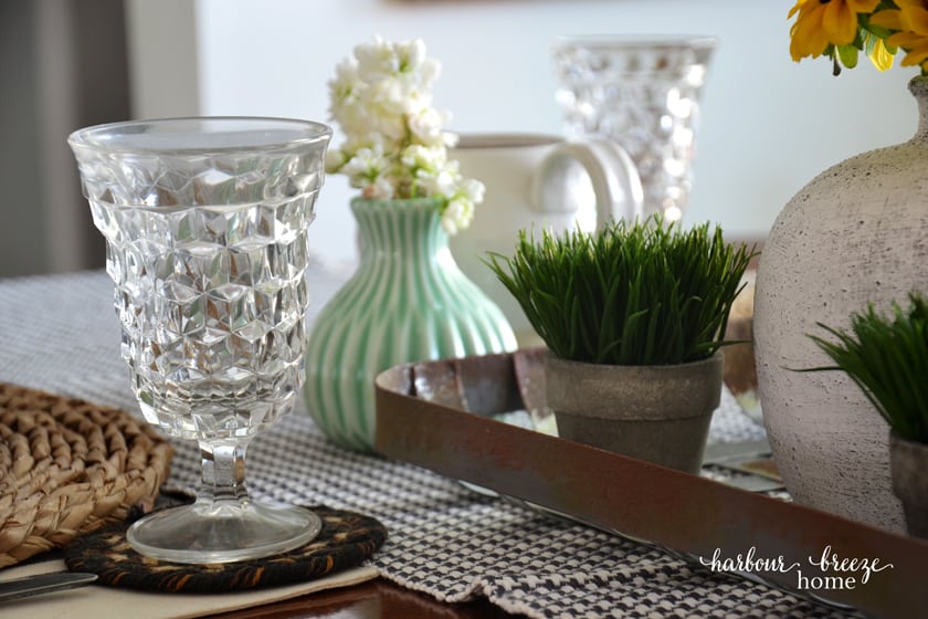 a clear glass with cut out feature set at the top of a table place setting with flowers nearby