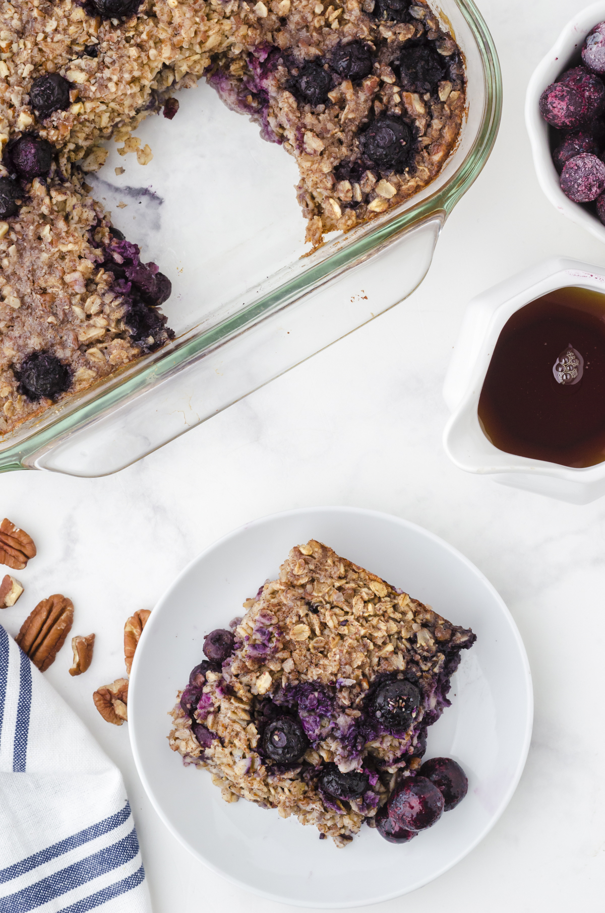 A slice of baked oats with bananas and blueberries on a serving plate with the pan of baked oats behind it.