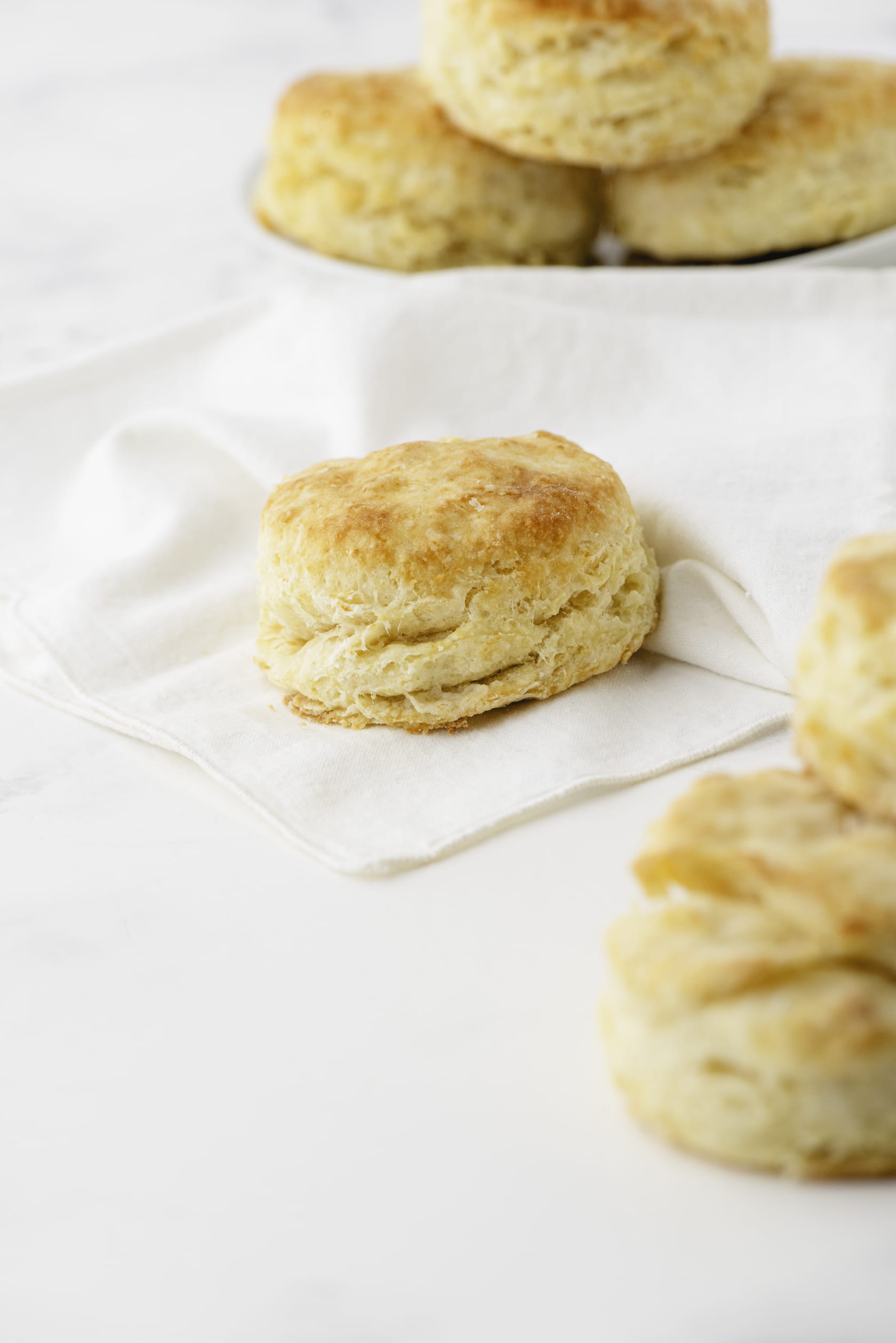 A flaky baking powder biscuit sitting on a cloth napkin with a plate of stacked biscuits behind it.
