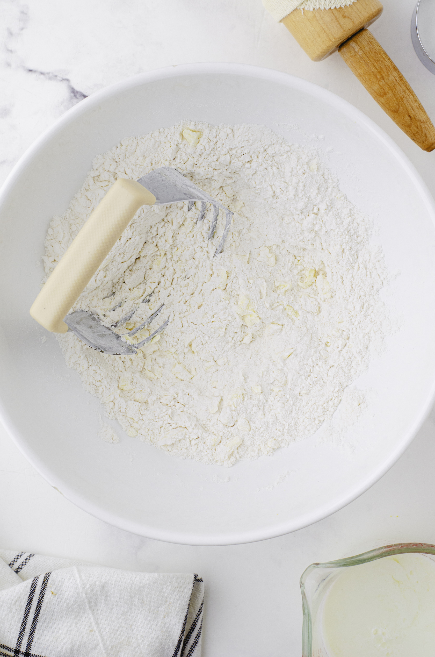 The dry ingredients for baking powder biscuits with butter that has been cut in in a large white mixing bowl with a pastry cutter sitting in it.