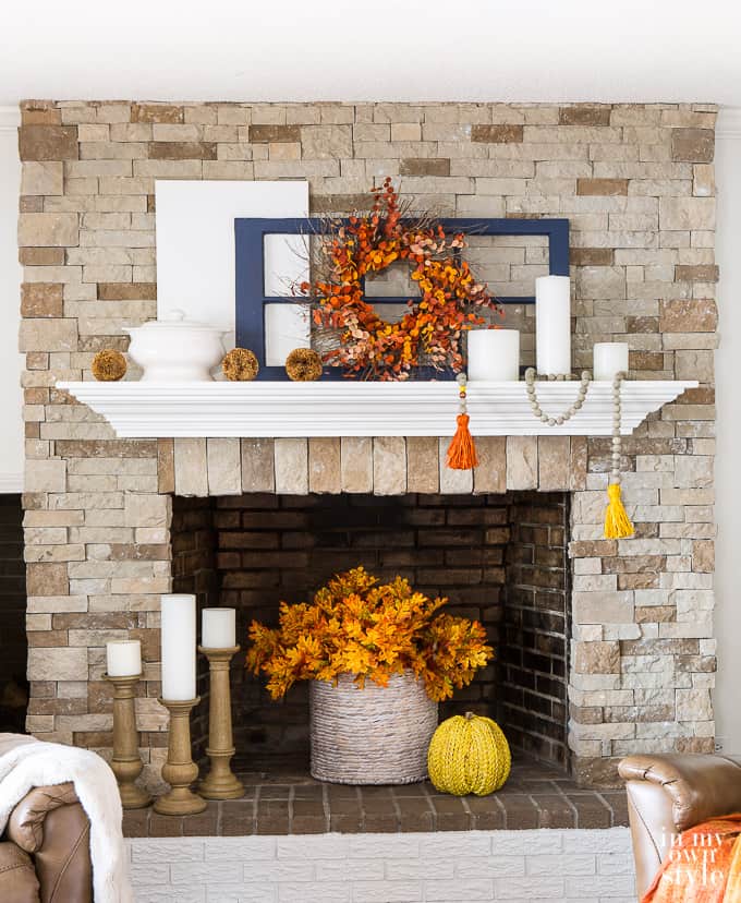 a fireplace mantel with a basket of fall leaves in the wood burning part and a blue window on the mantel with a fall wreath in front of it