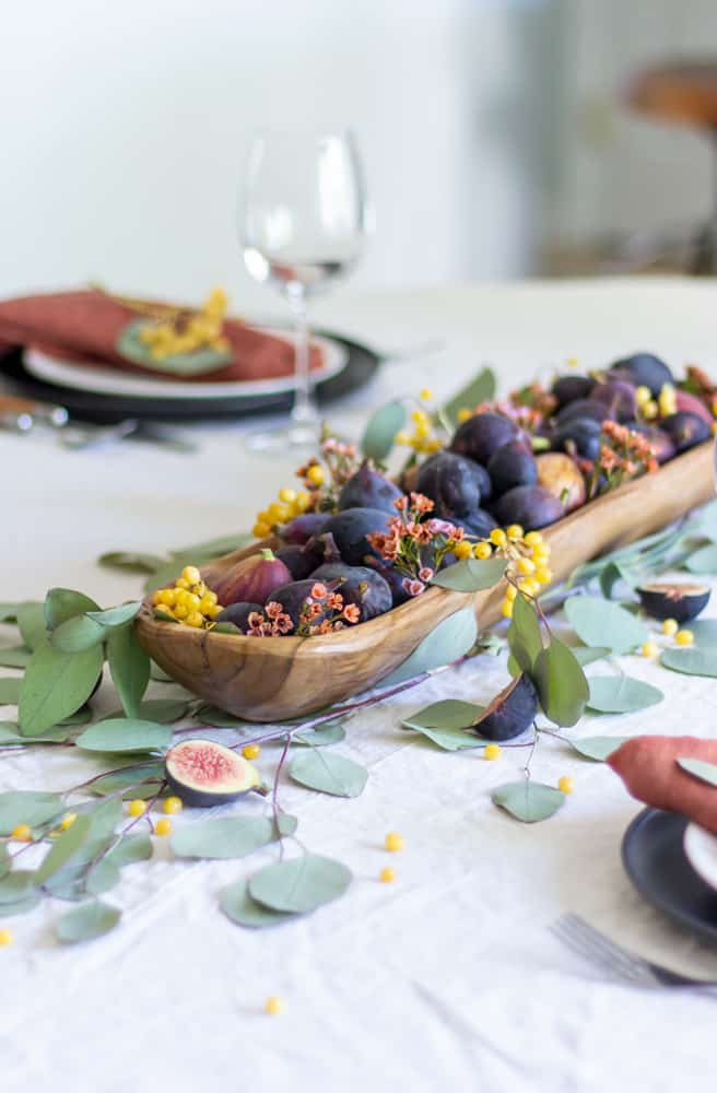 a table centerpiece made of a long wooden bowl filled with figs and faux greenery