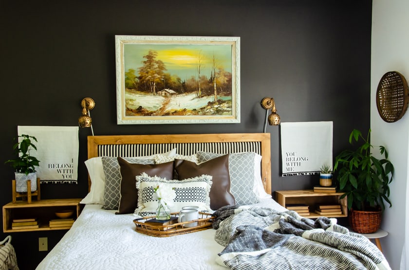 Boho style bedroom with the bed up against a black bedroom wall. A large vintage landscape art scene is centered above the headboard. Leather throw pillows are on the bed along with soft, woven, and cozy linens. Floating wooden nightstands flank the bed with plants and books as decor.