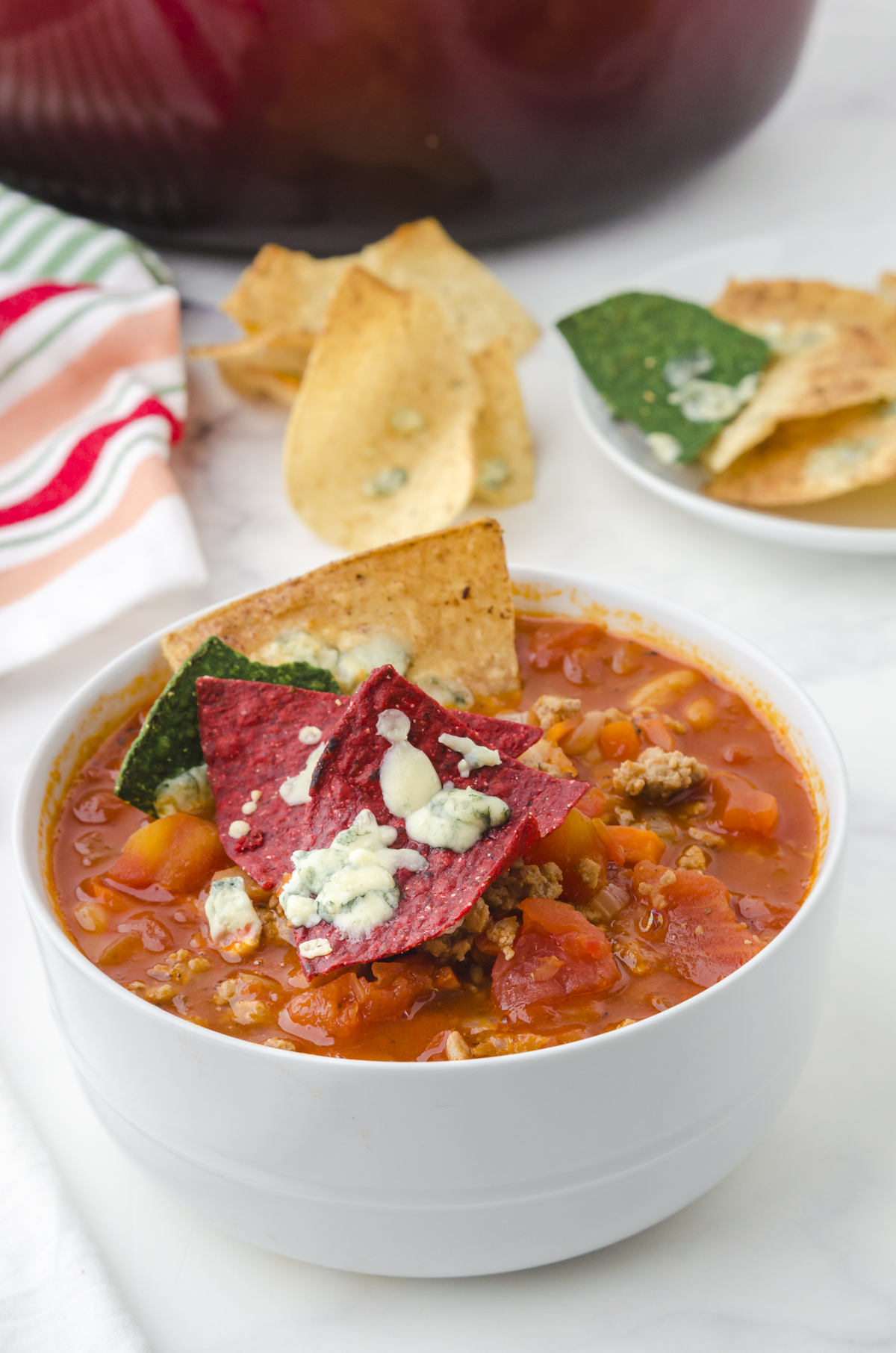 A close up of a bowl of buffalo chicken chili with red tortilla chips on the top with melted blue cheese crumbles.