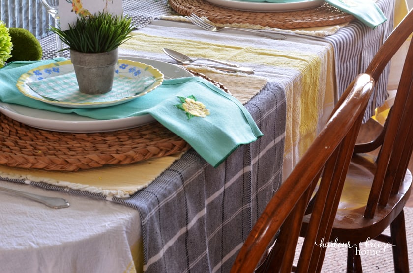 Layer the table with a yellow striped tablecloth, plaid dishtowels under the placesetting, and stacked plates on a woven charger.