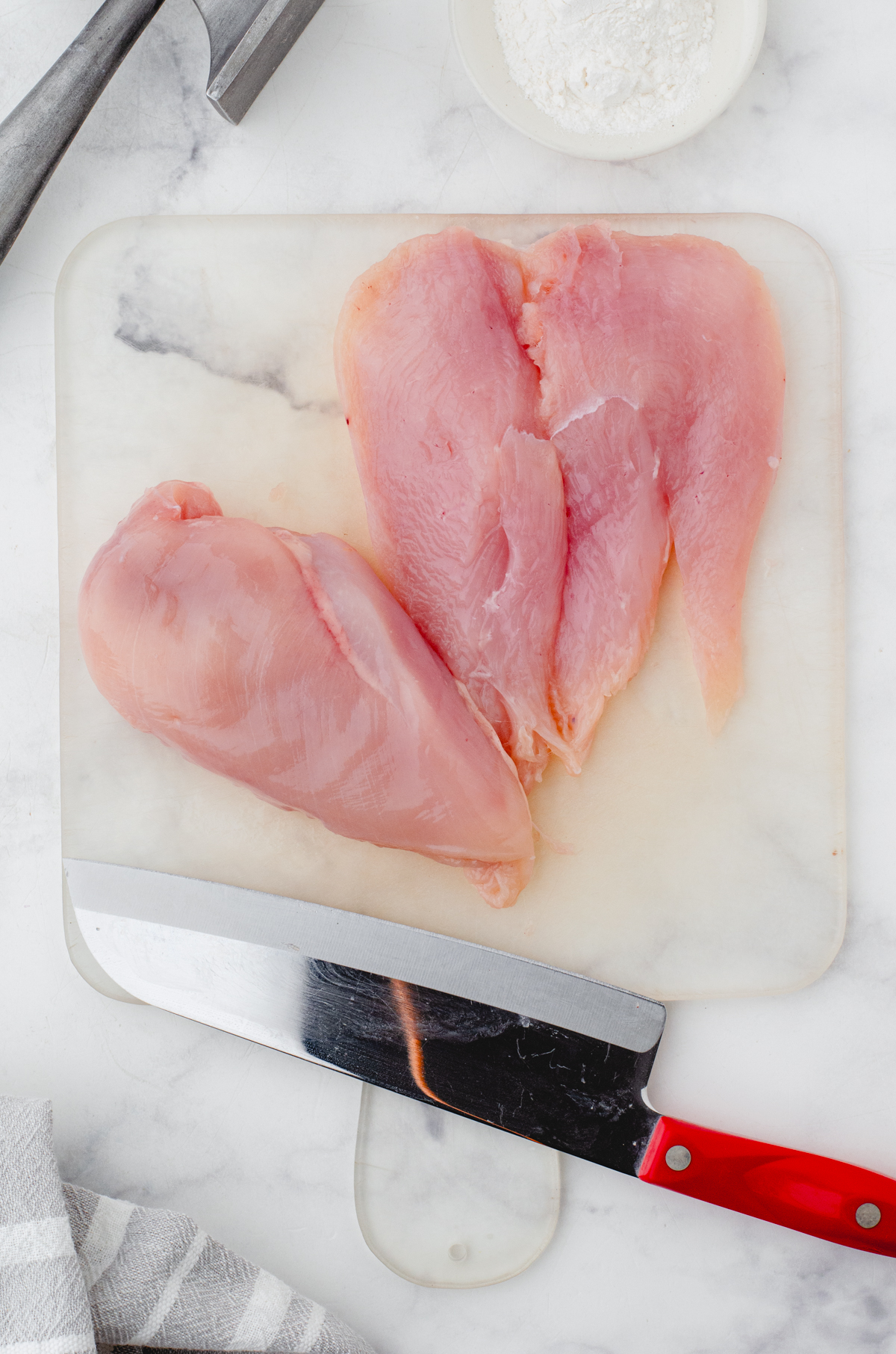 Chicken breasts on cutting board being butterflied with a large knife.