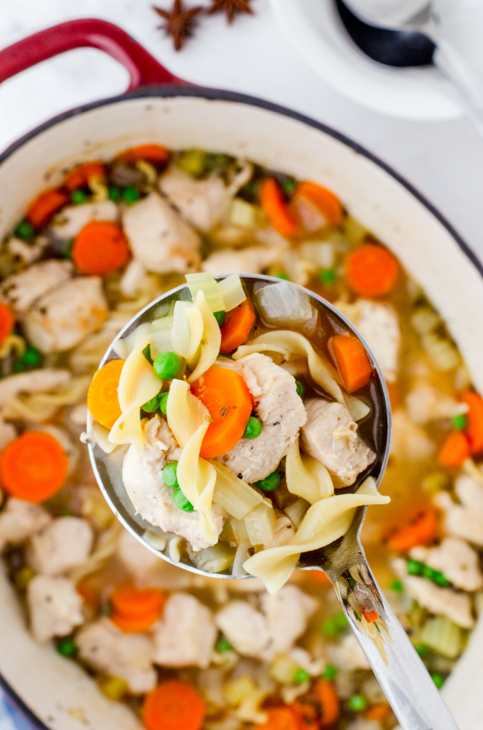 A soup ladle full of chicken noodle soup showing the bits of chicken, carrots, noddles, peas, and sauted onions.