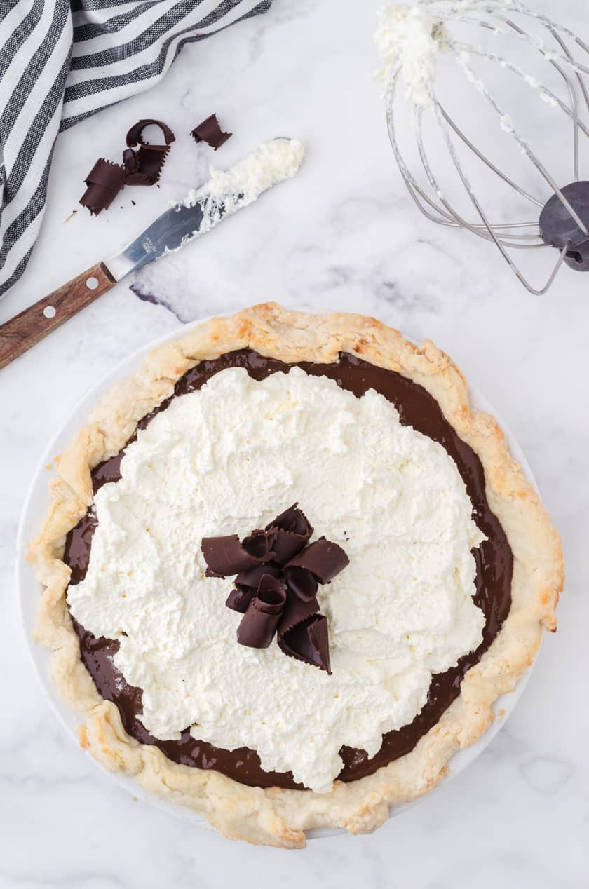 A whole chocolate pudding cream pie with a mixing bowl whisk with whipping cream beside.