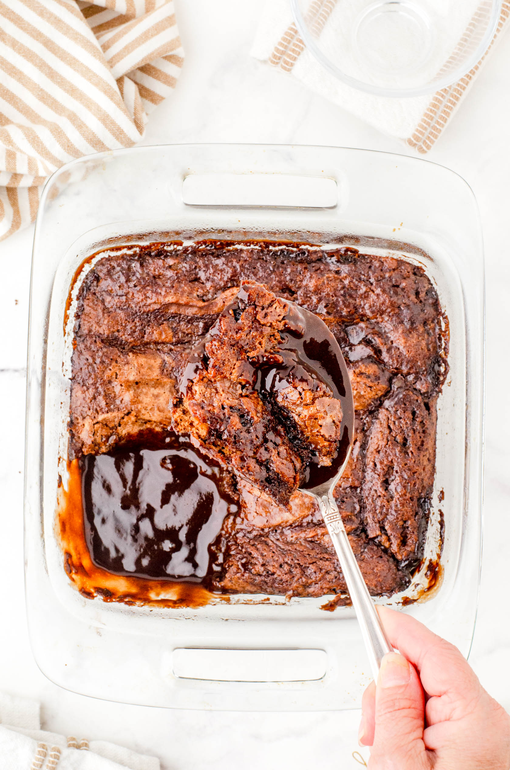 A scoop of hot fudge chocolate pudding cake being held above a pan of it showing how the cake and pudding are baked together.