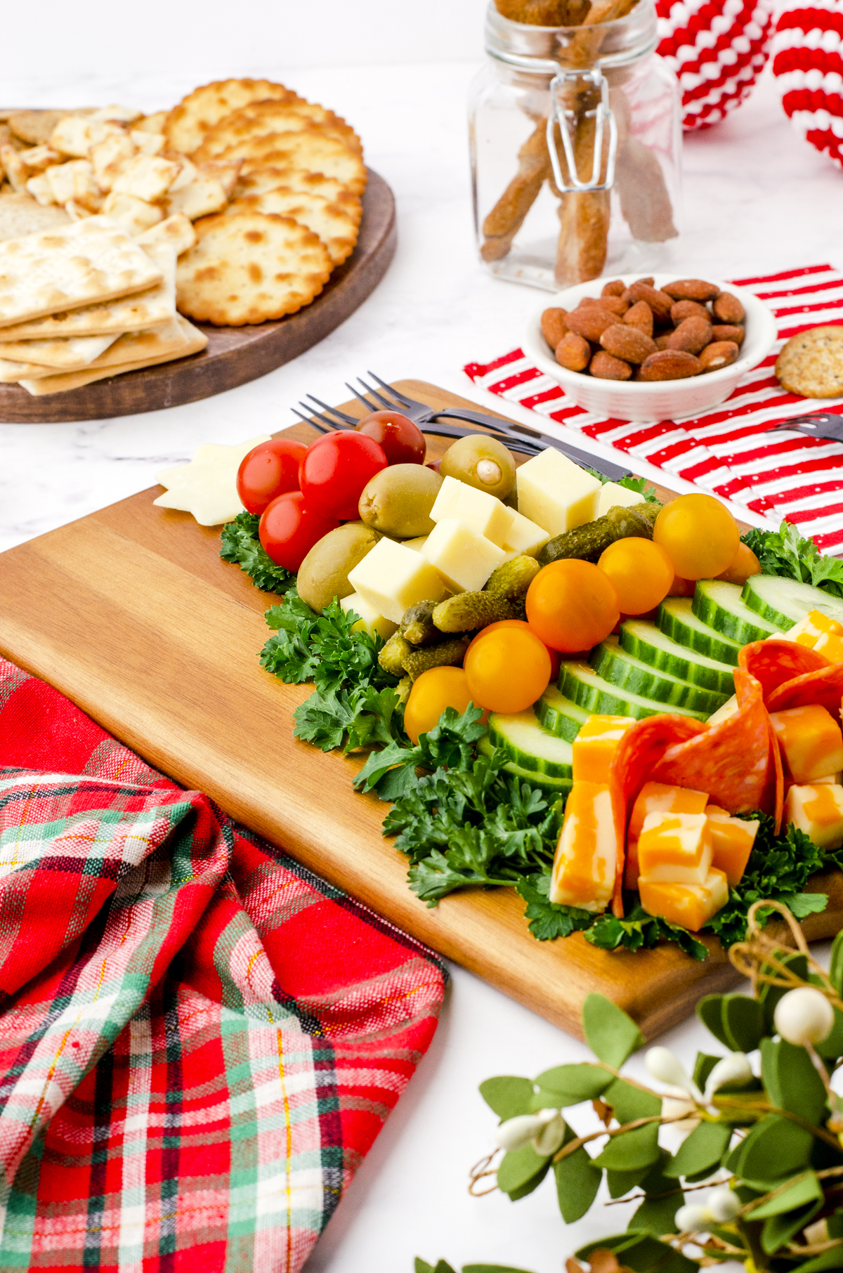 A christmas tree charcuterie board on a serving table surrounded by a platter of crackers, Christmas napkins, and a bowl of nuts.