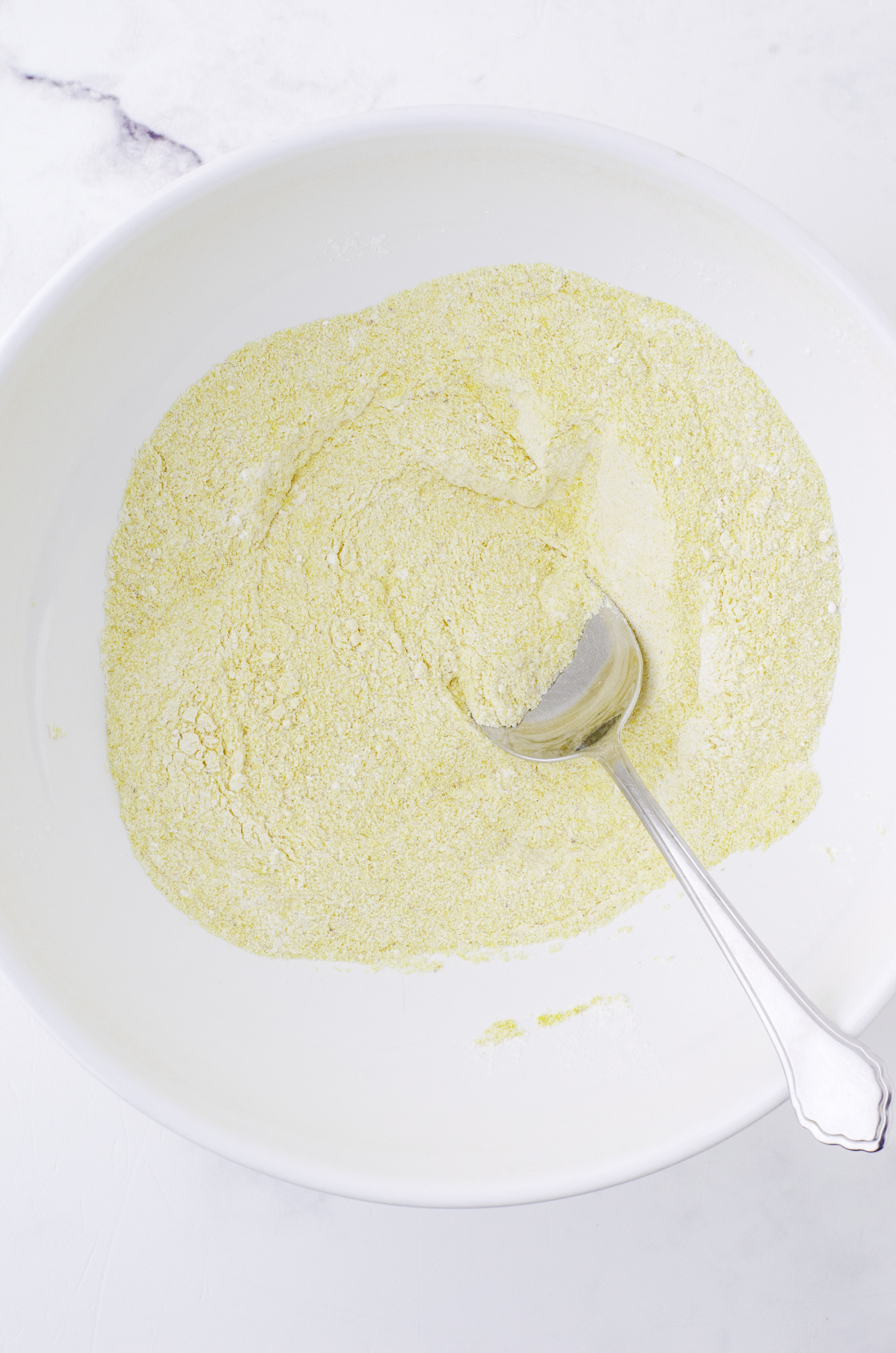 Dry ingredients for this cornbread recipe being stirred together in a large white mixing bowl with a big spoon.