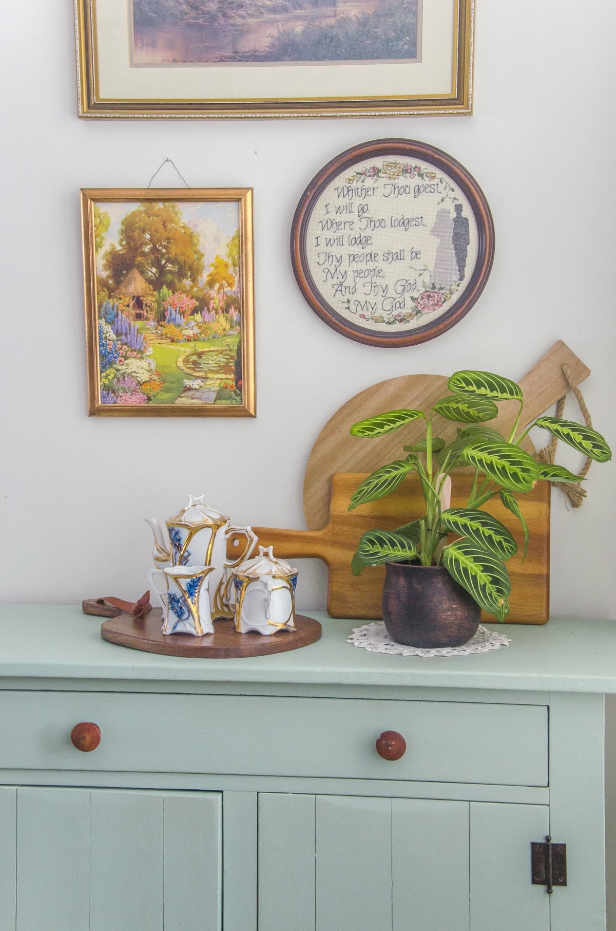 A cottagecore style wall art arrangement of vintage art hung above a dresser with a wooden tray and a plant on it.