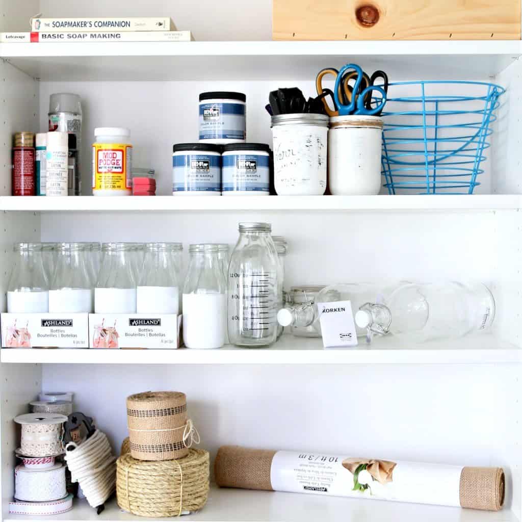 craft paint, scissors, mod podge, ribbons, craft books, and decorative jars organized on a white bookshelf