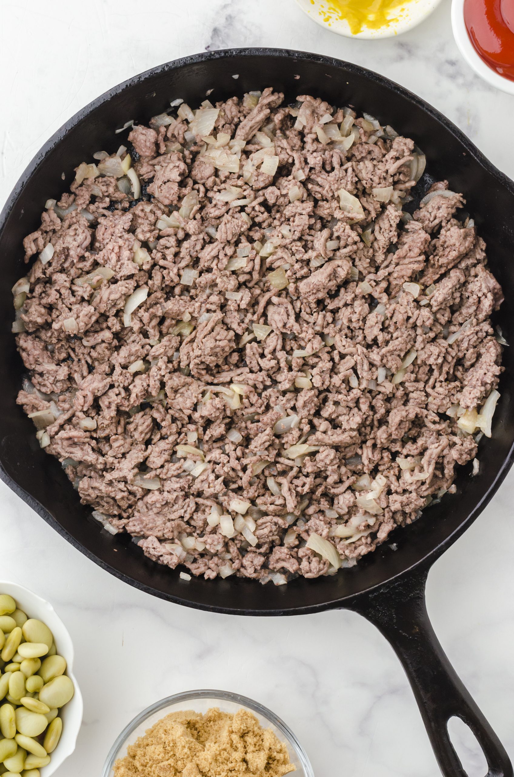 Browned ground beef and onions in a cast iron skillet ready to be put in a crock pot for this baked beans recipe.