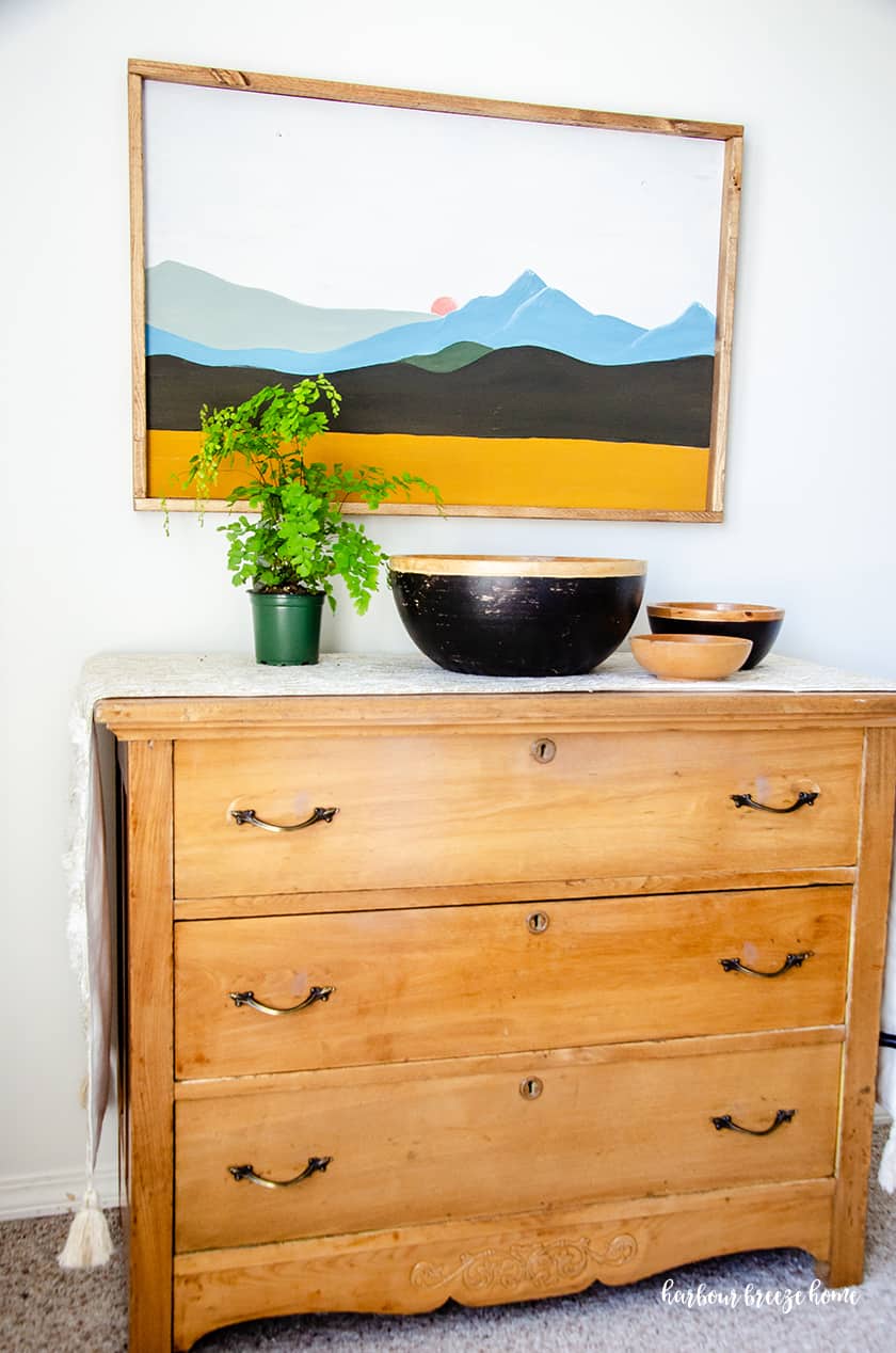 An easy mountina picture in a boho style hung above a wooden dresser with plants and wooden bowls in front of it.