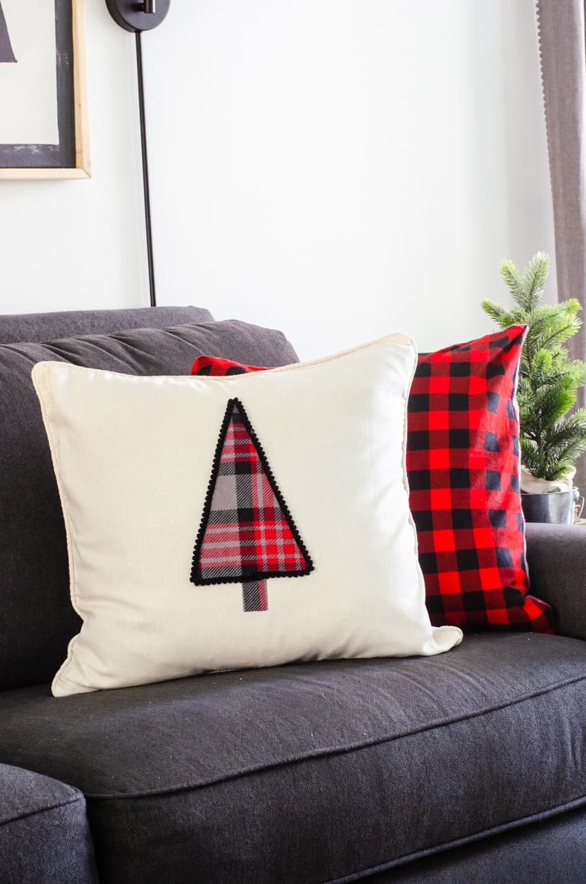 A DIY Christmas tree pillow propped up on a grey couch in front of a red buffalo check flannel pillow.
