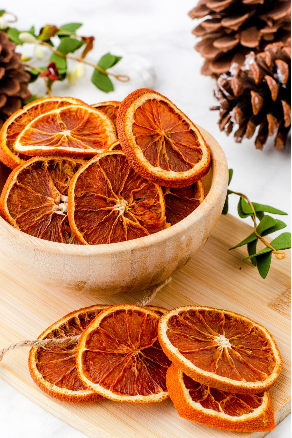 Dried orange slices in a wooden bowl sitting on a wood board surrounded by pine cones and faux greenery as an illustration of dried orange slices decor