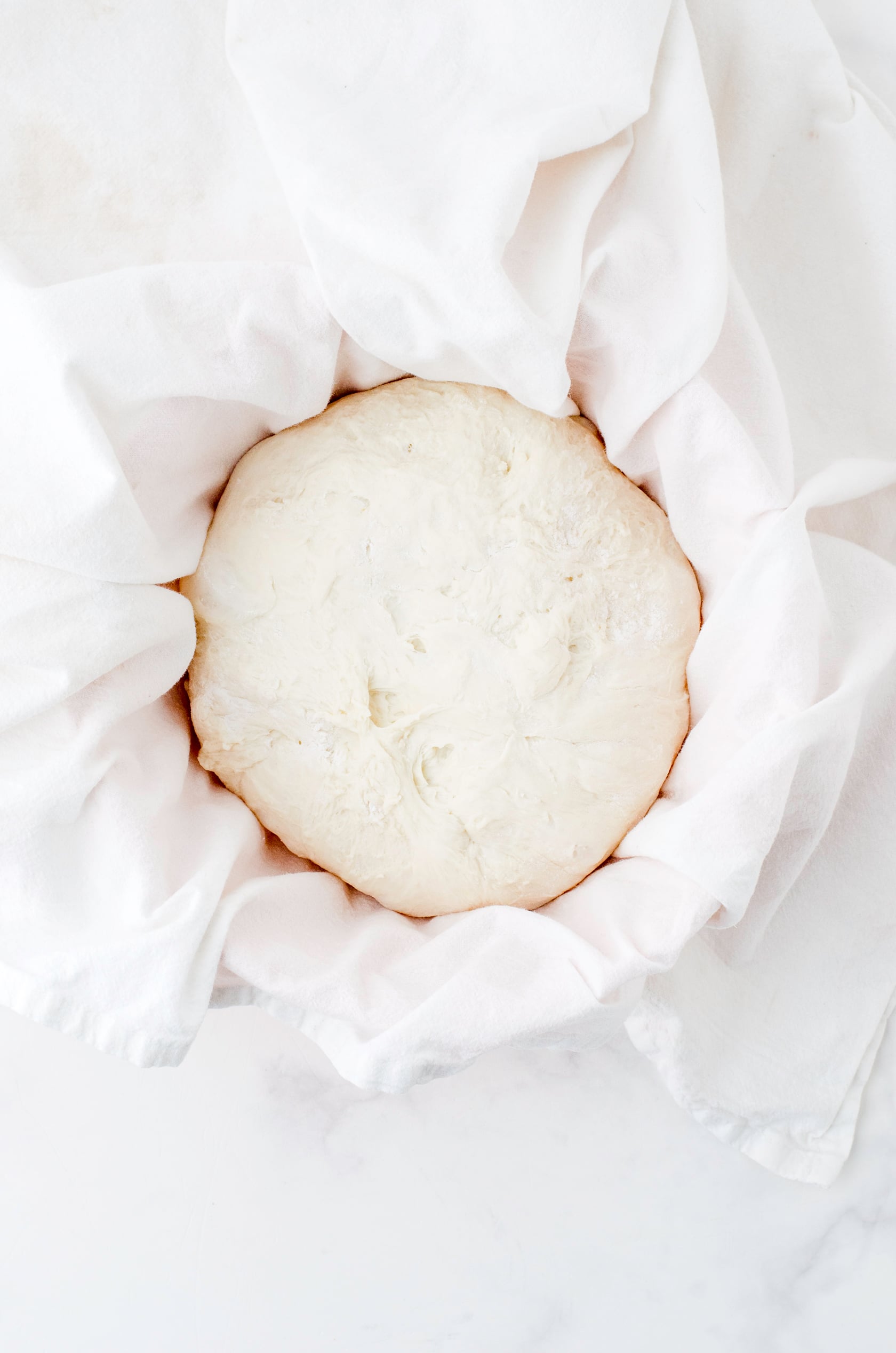 Sour dough bread formed and put in a bowl covered with a flour sprinkled dishtowel.