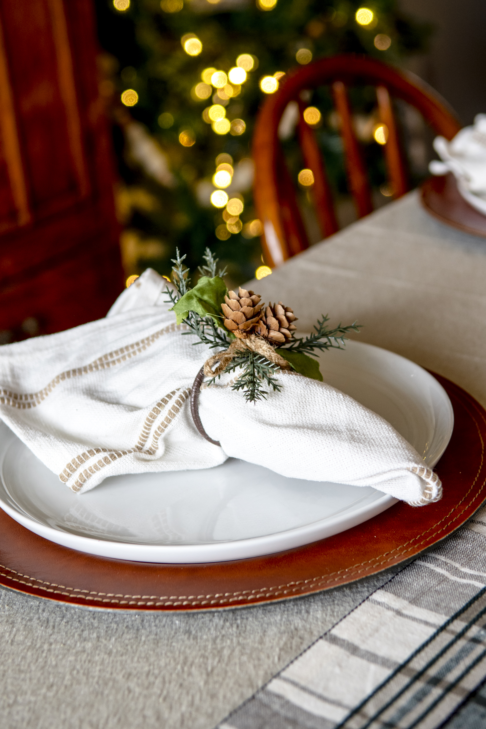 A leather charger plate with white plate on top and a cloth napkin with a napkin ring with pine cones on it.