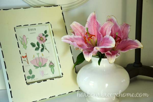 Pink lillies in a white vase sitting beside a framed botanical print
