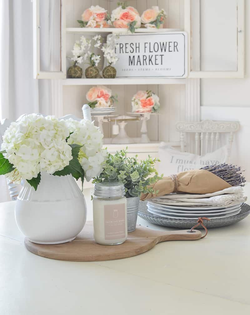 White Dining Room table with a flower centerpiece on it and a hutch in the background with coral flowers