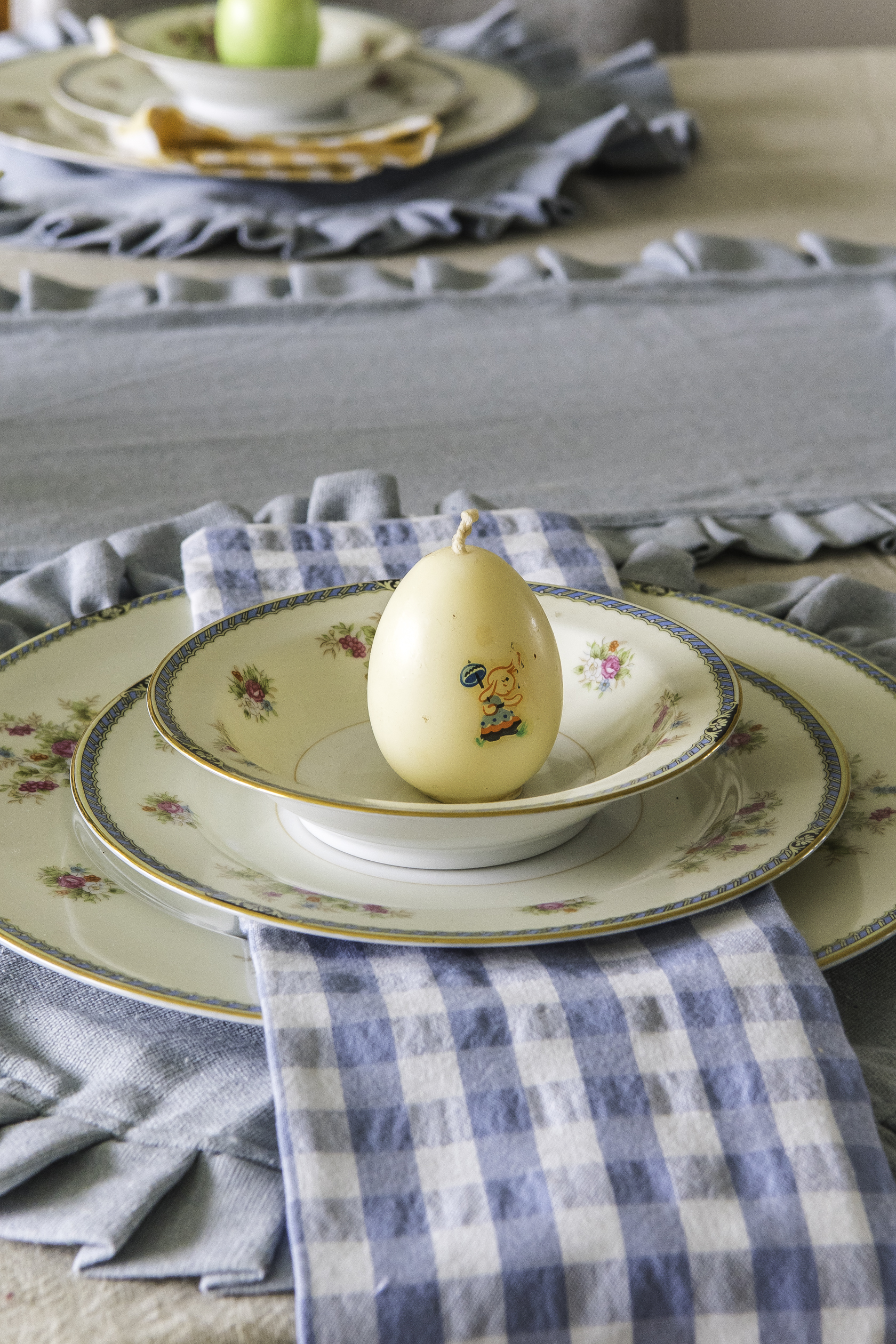 A table setting idea with a blue round ruffled placemat, blue and rose vintage china, blue checked cloth napkin, and a vintage egg candle sitting on the top of the top plate.