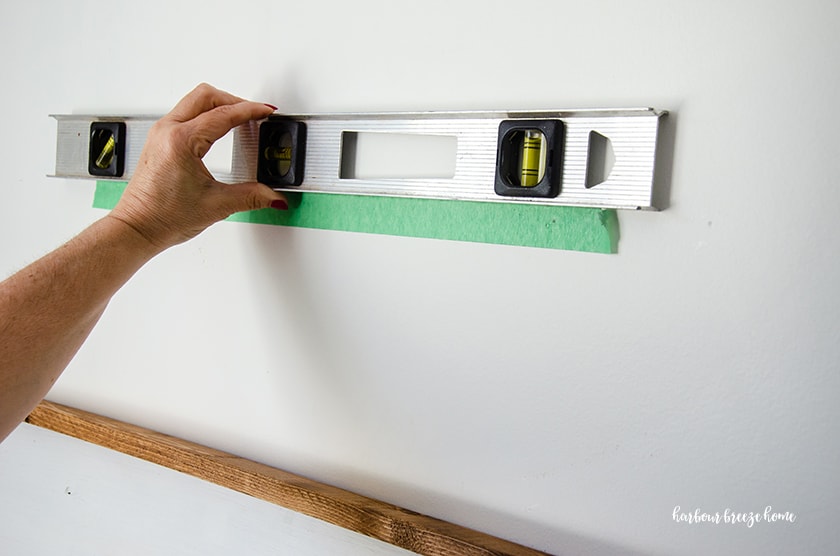 A woman holding a level at the top of a piece of painters tape with markings for hanging a picture with 2 sawtooth hangers.