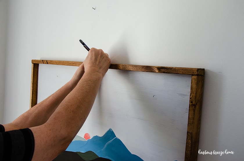 A picture being held up to a white wall with a person marking the center placement with a pencil.