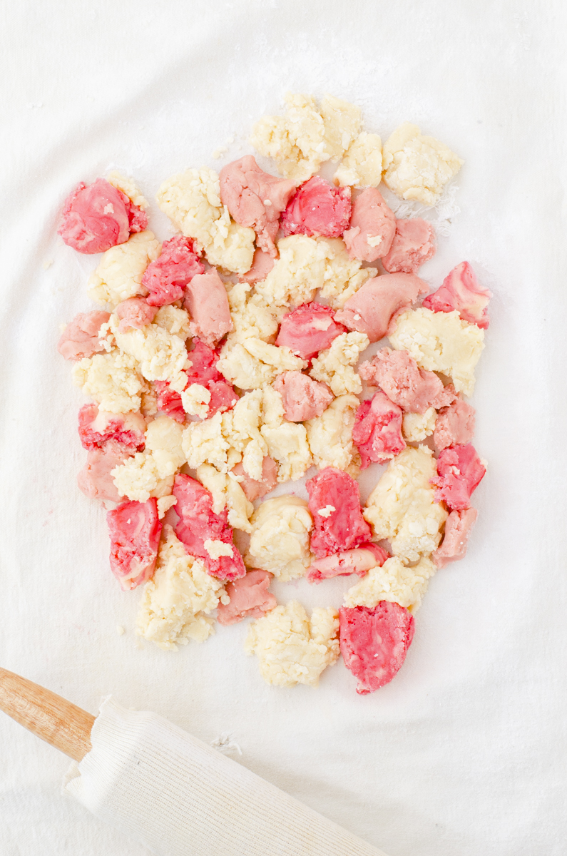Small chunks of different colored sugar cookies dough in alternating spaces on a pastry cloth ready to be rolled together.