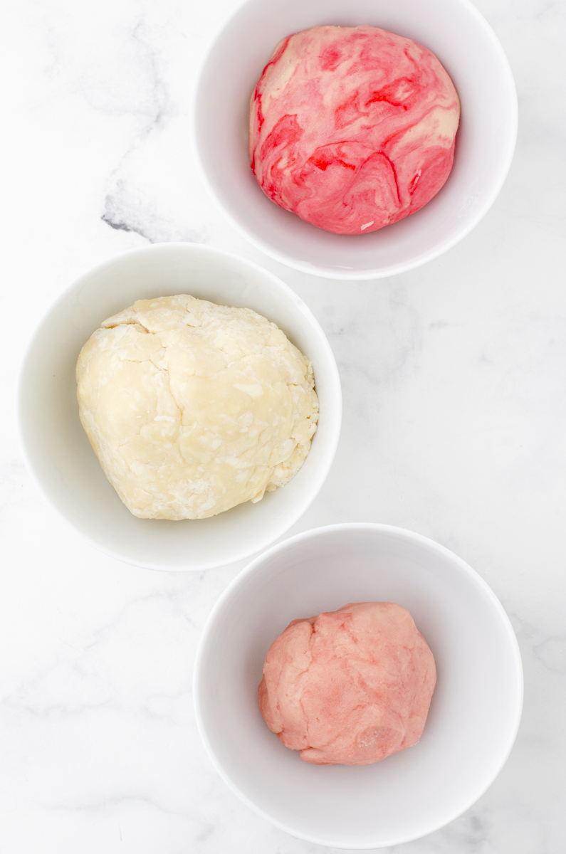 Sugar cookies dough separated into 3 dough balls. One is colored red marble, one is white, and one is pink.