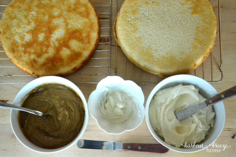 The parts of a stacked cake sitting on a table top. It shows 2 vanilla cake layers, a bowl butterscotch filling, bowls of icing, and 2 spreading spatulas.