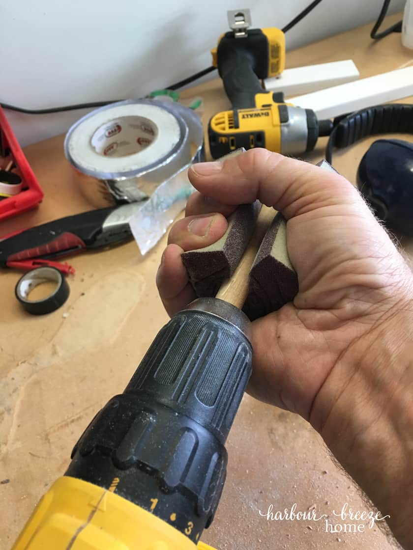 A dowel attached to the end of a drill with a sanding sponge squeezed around it with a man's hand.