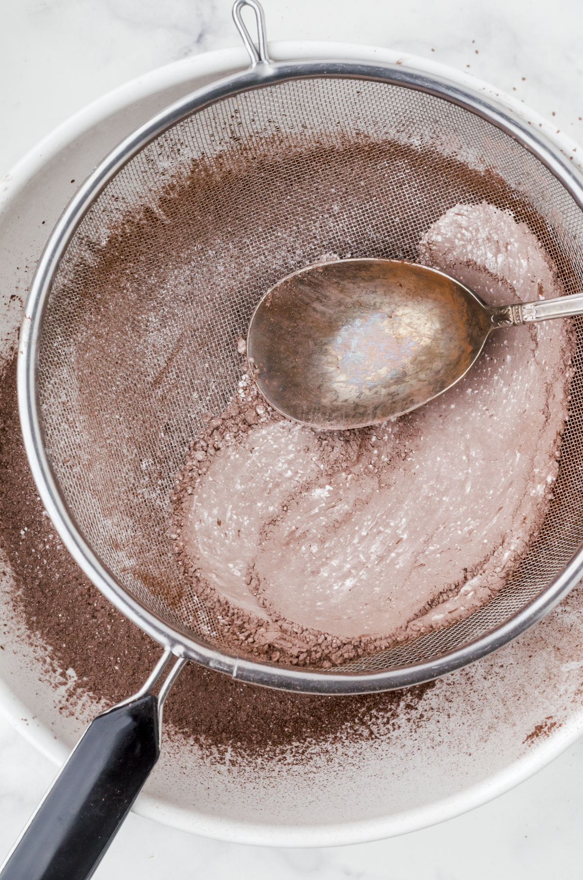A large spoon gently pressing out the lumps of cocoa powder and flower in the bottom of a fine mesh strainer.