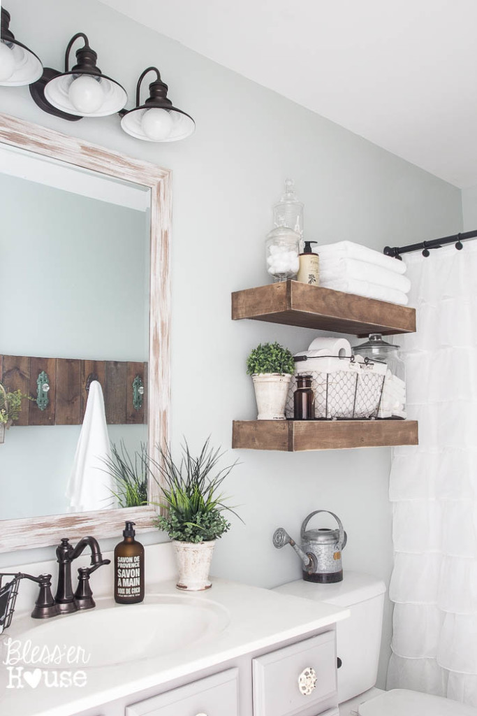 A bathroom design with farmhouse style similar to joanna gaines bathroom. It has 2 wooden shelves above the toilet, farmhouse style lighting about the vanity, and accessories like plants, wire baskets, and glass jars filled with cotton balls.