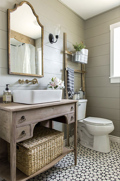 A bathroom design in the farmhouse style similar to what you would see on a Joanna Gaines Fixer Upper show. It has shiplap on the walls, a wooden framed mirror, and a ladder propped behind the toilet with a basket filled with towels attached.