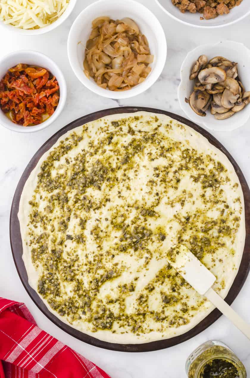 pesto sauce being spread over pizza dough on a stoneware pan
