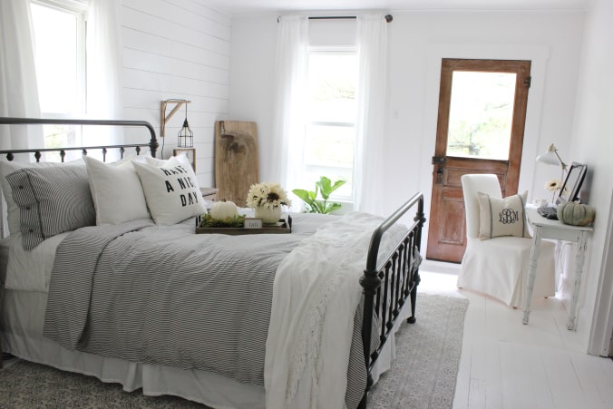 A white farmhouse bedroom with a metal bedframe bed in black, a small table for a desk and a wooden door leading outside.