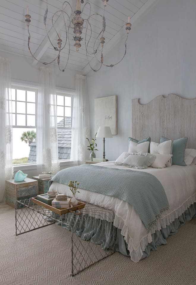 A farmhouse bedroom with a vaulted ceiling, large chandilier, and a bed with a white washed headboard, & white bedding with light blue accents.