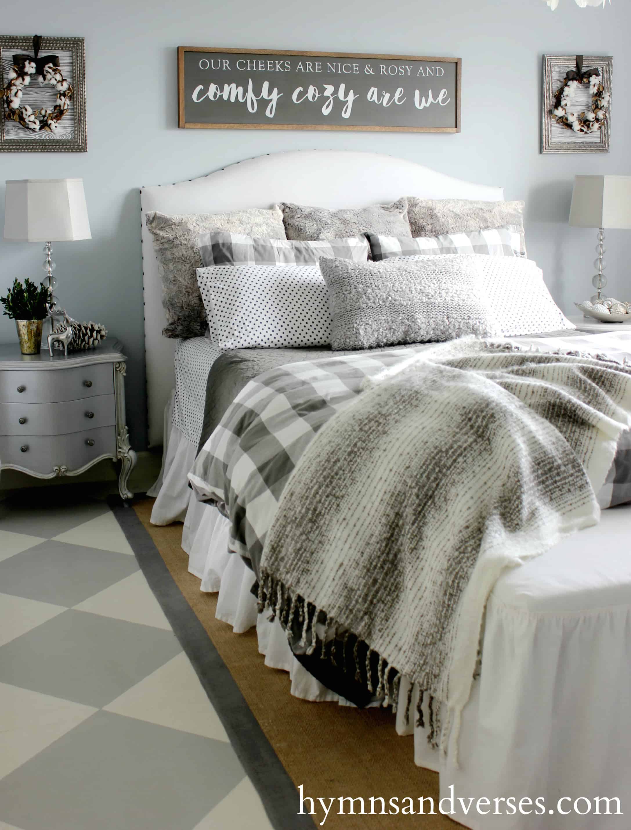 Farmhouse bedroom with white upholstered headboard on bed; a light blue wall behind, and gray bedding and pillows.