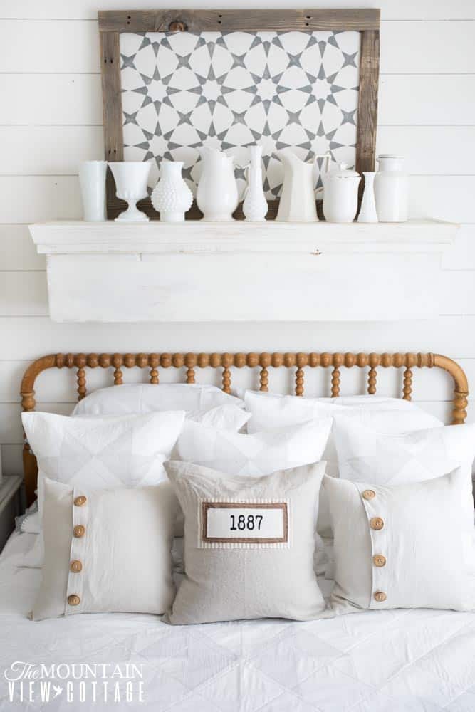 A farmhouse bedfroom with a wooden bedframe with a shelf above it with white pitchers and a sign.