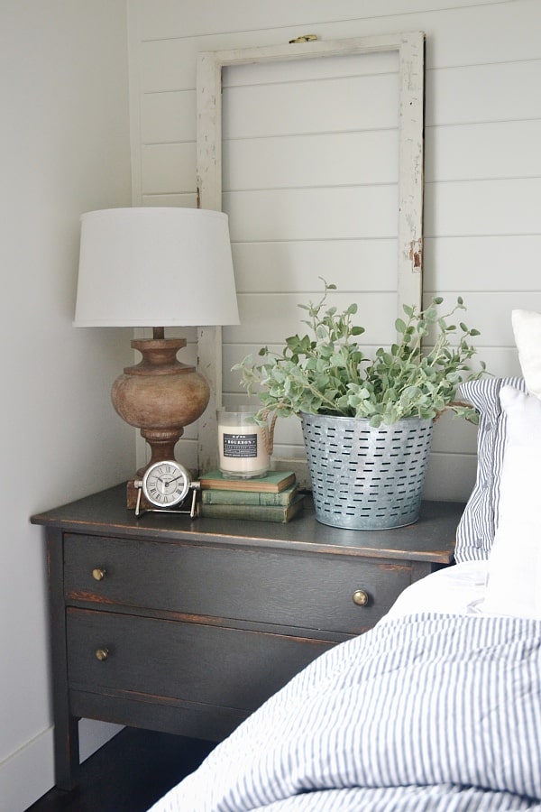 A farmhouse style nightstand beside a bed with an old window frame propped up on it and a galvanized bucket of greenery, stack of books, candle and lamp on it.