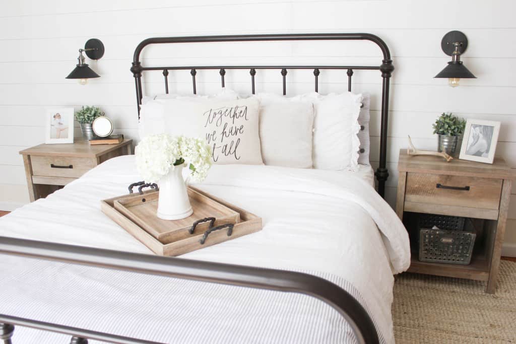 Farmhouse bedroom with metal bedframe, white shiplap wall behind, wood toned nightstands beside, and black wall mounted sconce lights above the nightstands.