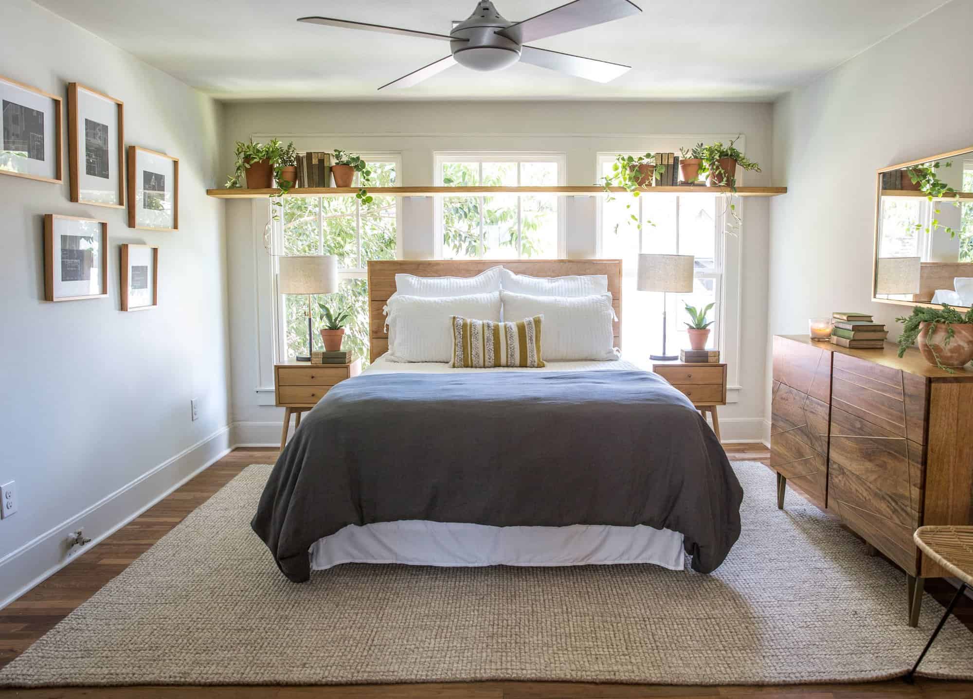 A wooden headboard on a bed in front of 3 windows with a long shelf above it with plants on it.