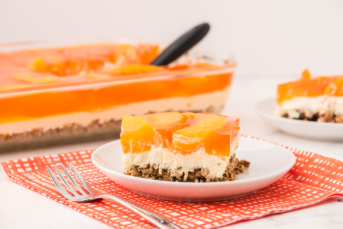 A slice of peach pretzel salad on a serving plate with the pan of peach pretzel salad behind.