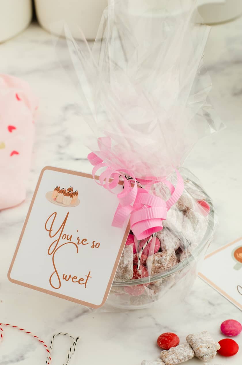 A clear bowl filled with valentines puppy chow mix with clear cellophane wrapped around it and a valentines tag that says "you're so sweet" tied to it.